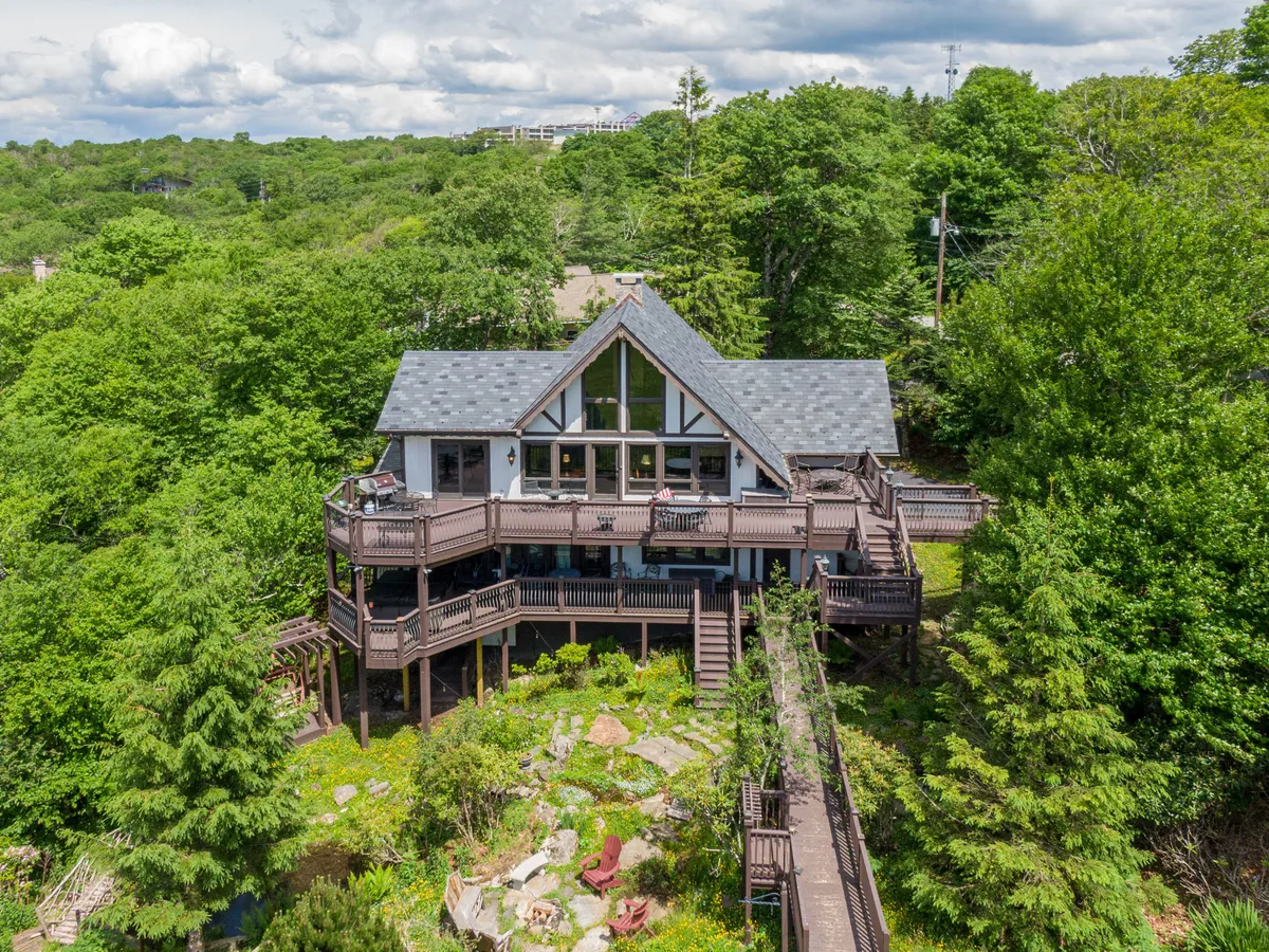 The hero image showcases the exterior of a spacious mountain home with expansive wrap-around decks, large windows that frame scenic mountain views, and natural landscaping that blends seamlessly into the serene surroundings. The deck is inviting for stargazing, morning coffee, and sunset dining, offering multiple outdoor spaces for relaxation and enjoying the beauty of Beech Mountain.