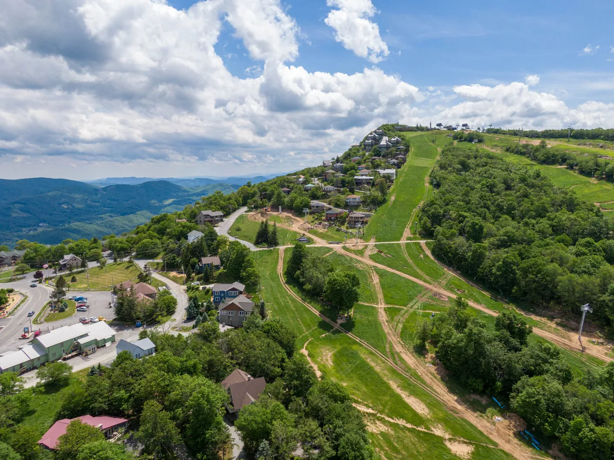 A wide view of rolling hills, scattered houses, winding roads, and lush greenery. The landscape hints at year-round activities: hiking, biking, and skiing, with an adventurous and serene mountain vibe.