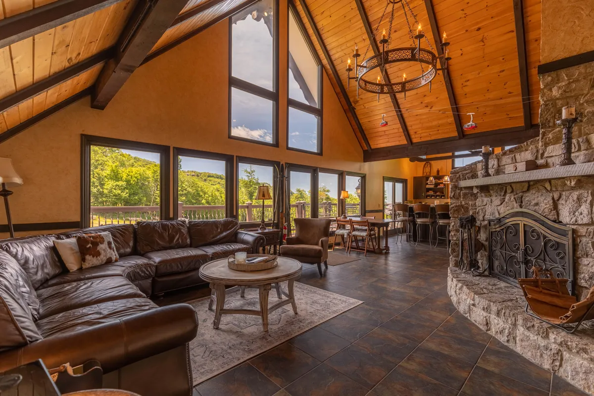 A spacious great room featuring large windows that let in natural light, a striking stone fireplace, a leather sectional sofa, and exposed wooden ceiling beams. The arrangement is inviting and set up for gatherings, with comfortable seating oriented toward the fireplace and windows framing scenic views.