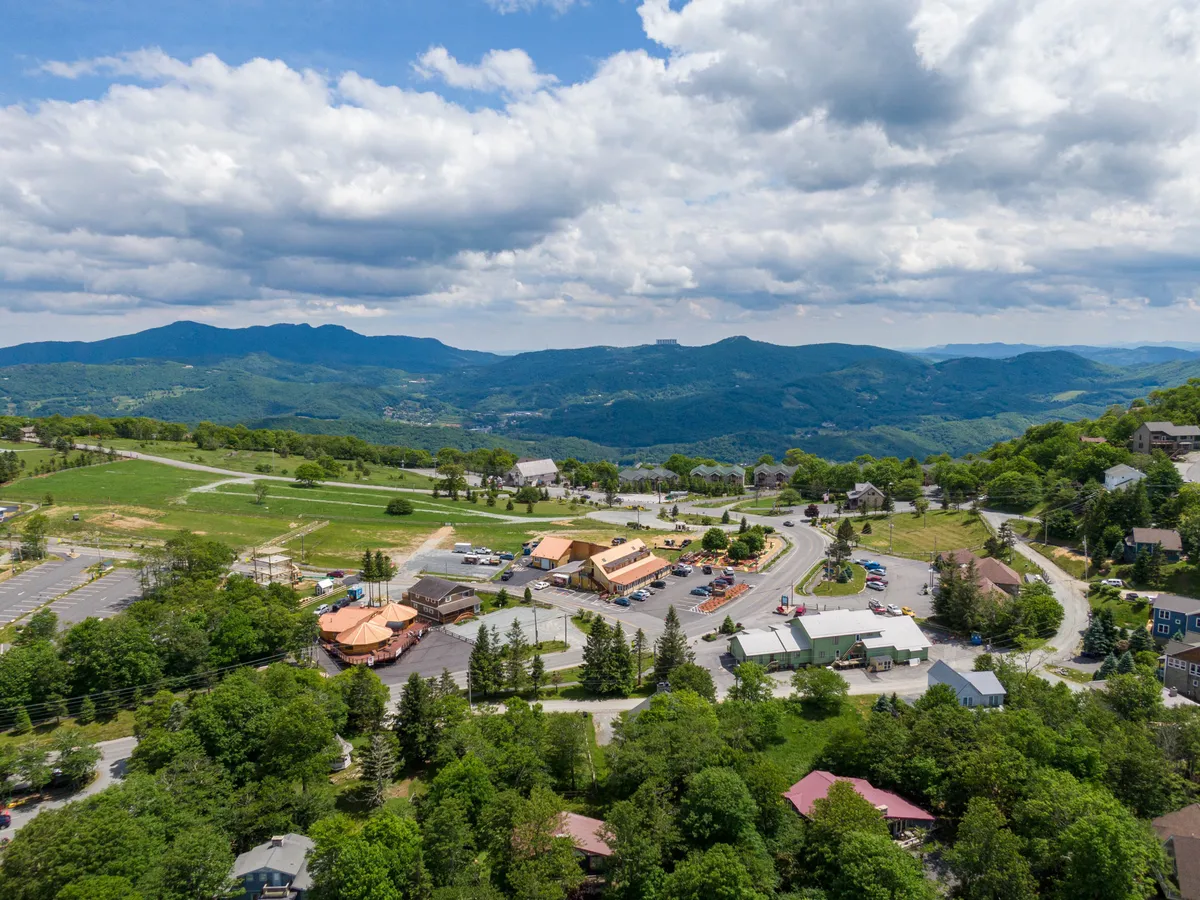 A panoramic view of Beech Mountain displays a mountain range, local residential buildings, commercial establishments, and a winding road. Lush green trees fill the scene, conveying the vibrancy of the area and its walkable access to local shops and attractions.