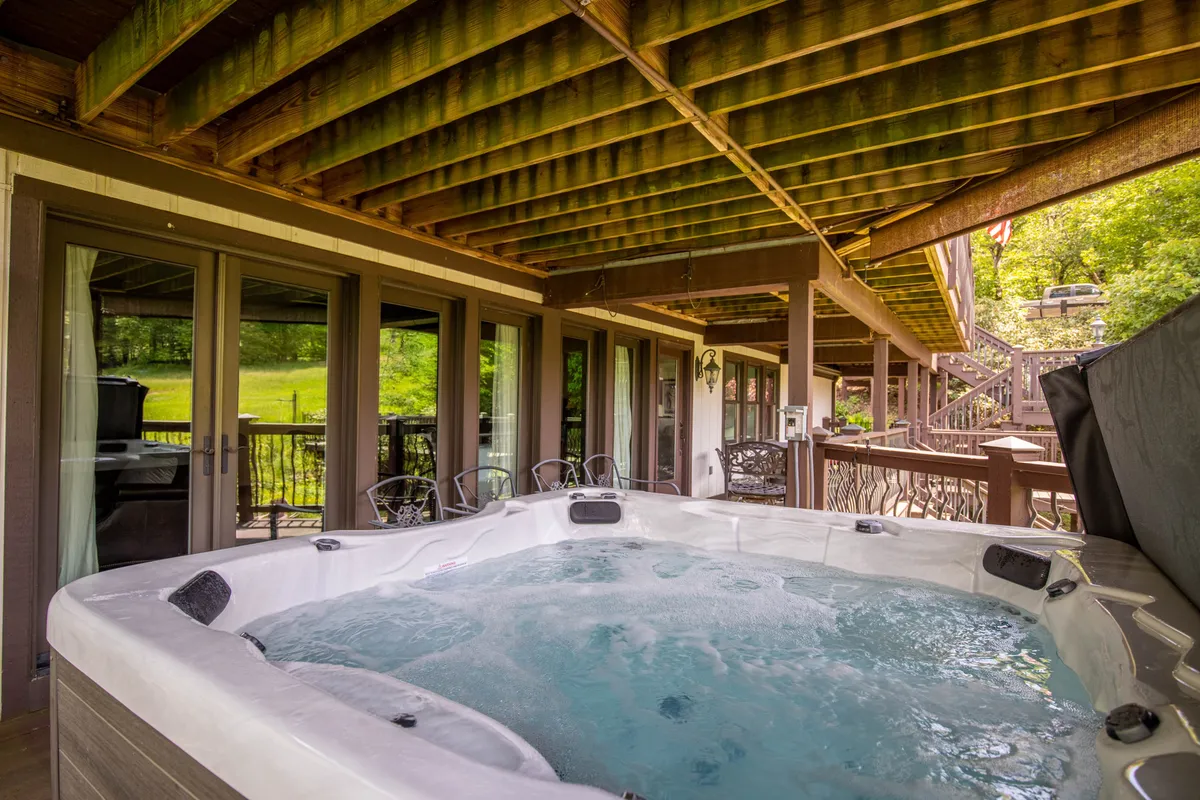 A tranquil scene featuring a hot tub set on a spacious wooden deck, with glass doors connecting the interior to the exterior. The surroundings suggest privacy and offer a sense of serenity, perfect for relaxation or socializing while enjoying the mountain air.