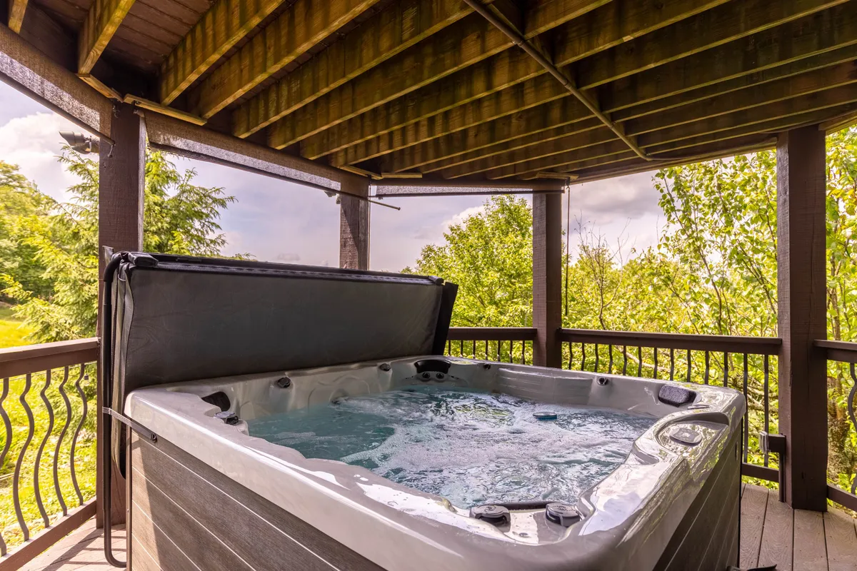 A large outdoor hot tub is set on a wooden deck surrounded by lush greenery and trees. The deck's railing frames the scene, creating a private, tranquil atmosphere perfect for soaking and socializing while enjoying the view.