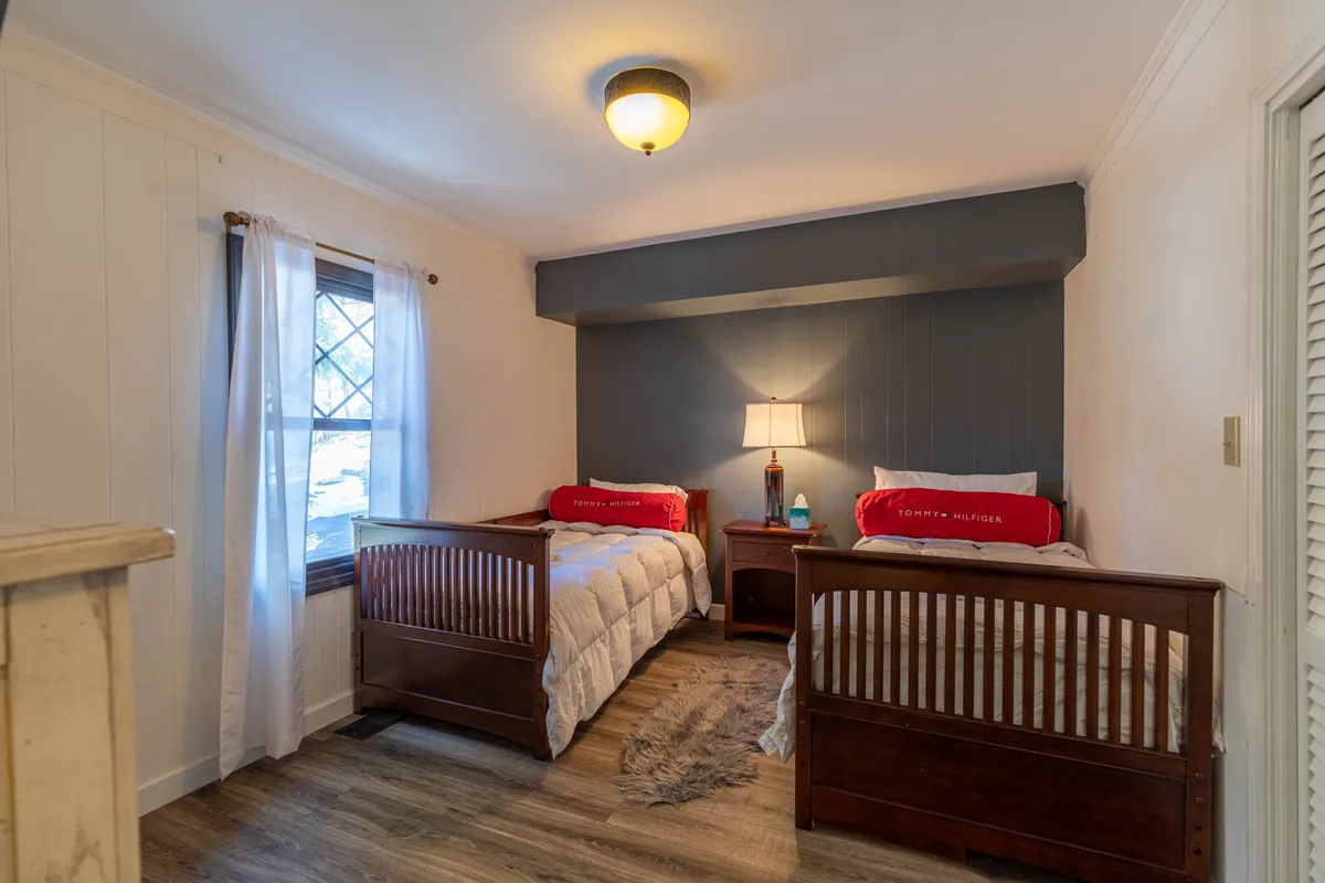 A bright, inviting bedroom with two twin beds dressed in red pillows, a central nightstand with a lamp, and a large window with sheer curtains. The setting is cozy, ideal for kids or additional guests, and radiates warmth and playfulness.