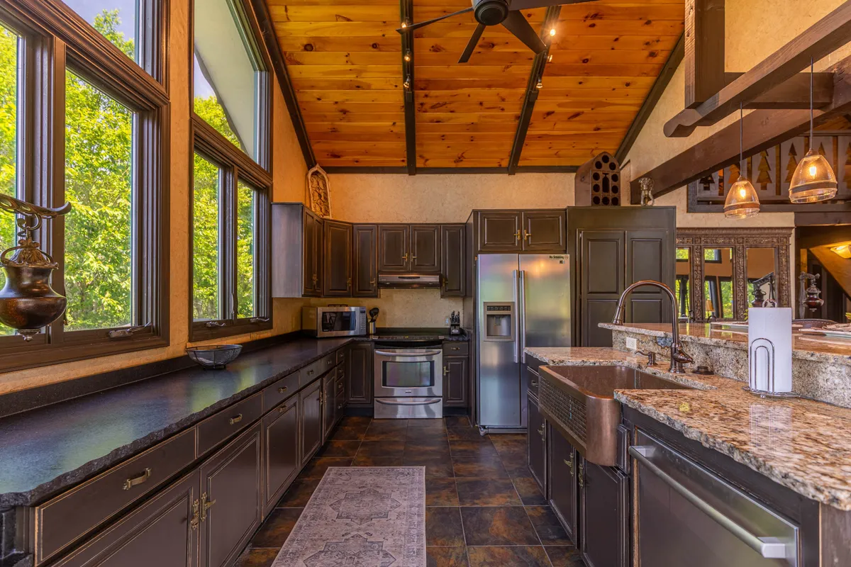 A modern kitchen with granite countertops, stainless steel appliances, dark cabinetry, wooden ceiling accents, and large windows. The kitchen is open and seamlessly connected to the main living space, creating an ideal setup for entertaining and group meals.