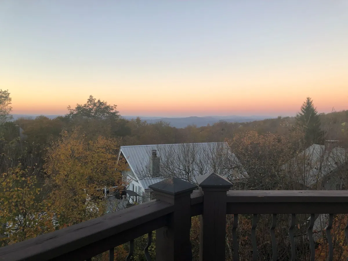 A breathtaking view of distant mountains beneath a sunset sky, framed by autumn foliage and a wooden railing. The vantage point is perfect for quiet moments of reflection, evening drinks, or simply soaking in the stunning natural surroundings.