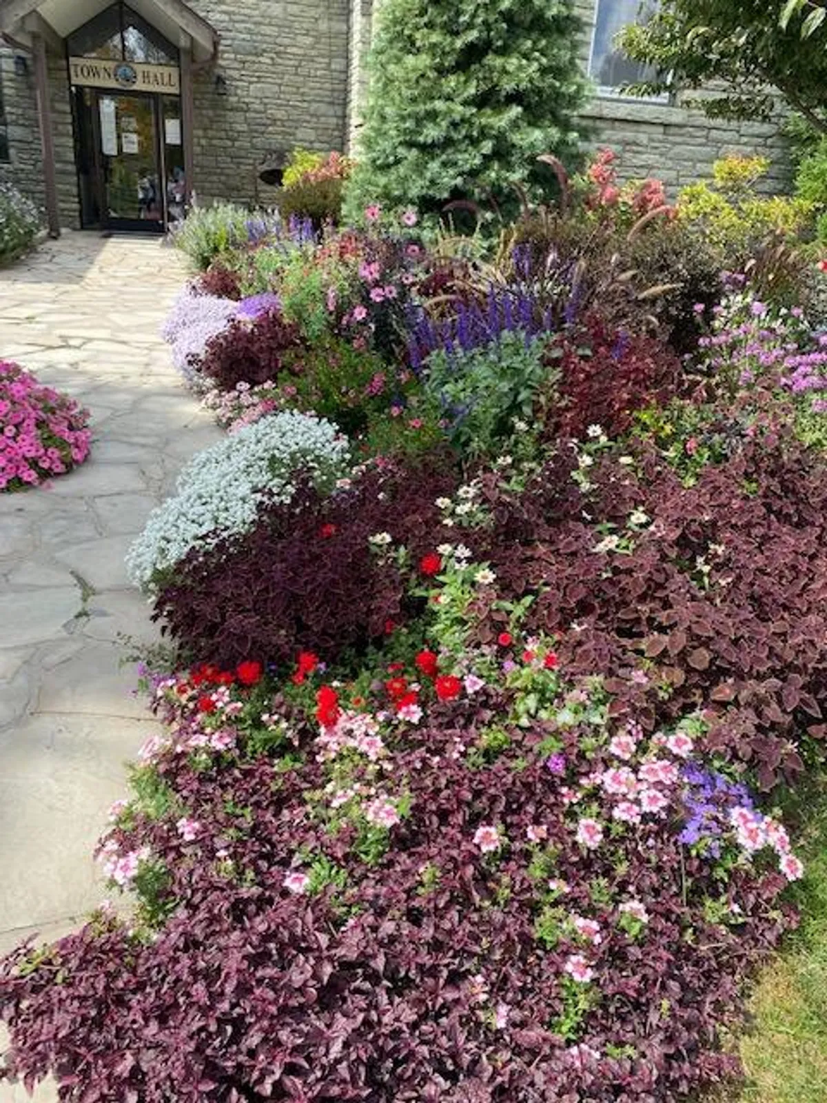 A vibrant exterior shot of Beech Mountain's Town Hall, surrounded by colorful, seasonal flower beds and a stone pathway leading to the entrance. The scene suggests a welcoming community hub where local events and public gatherings take place.