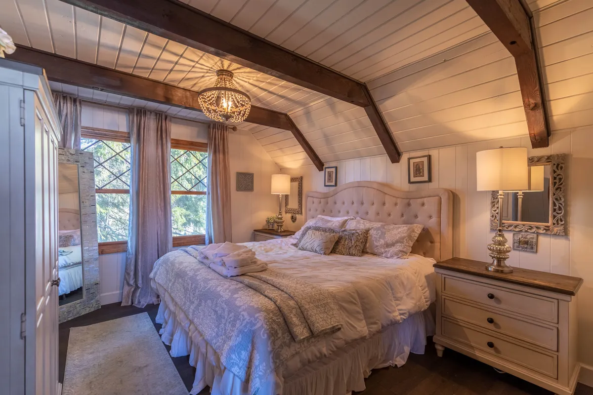 A tranquil bedroom scene with a plush bed adorned with decorative pillows, an elegant chandelier overhead, a mirrored wardrobe, and a window framed with gentle curtains. Soft natural light streams in, enhancing the serene atmosphere.