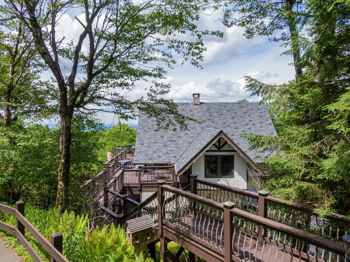 This exterior shot showcases the house with its wooden deck set amid lush trees, emphasizing the property's natural surroundings and its proximity to trails and outdoor adventure. The visible deck invites viewers to imagine relaxing after a day on the slopes or trails.