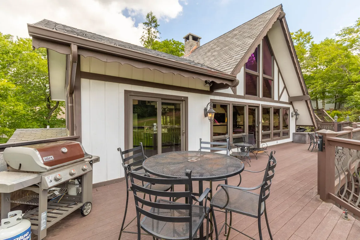 An expansive outdoor dining area is set on a spacious deck, featuring a large table with chairs, a grill for barbecuing, and large windows connecting the space to the interior of the house. The surrounding greenery provides a peaceful backdrop, and the setup invites social gatherings and relaxed meals.